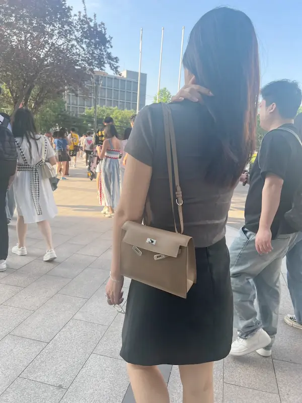 Woman wearing Etoupe Grey Replica Hermes Kelly Danse crossbody with coffee in city setting